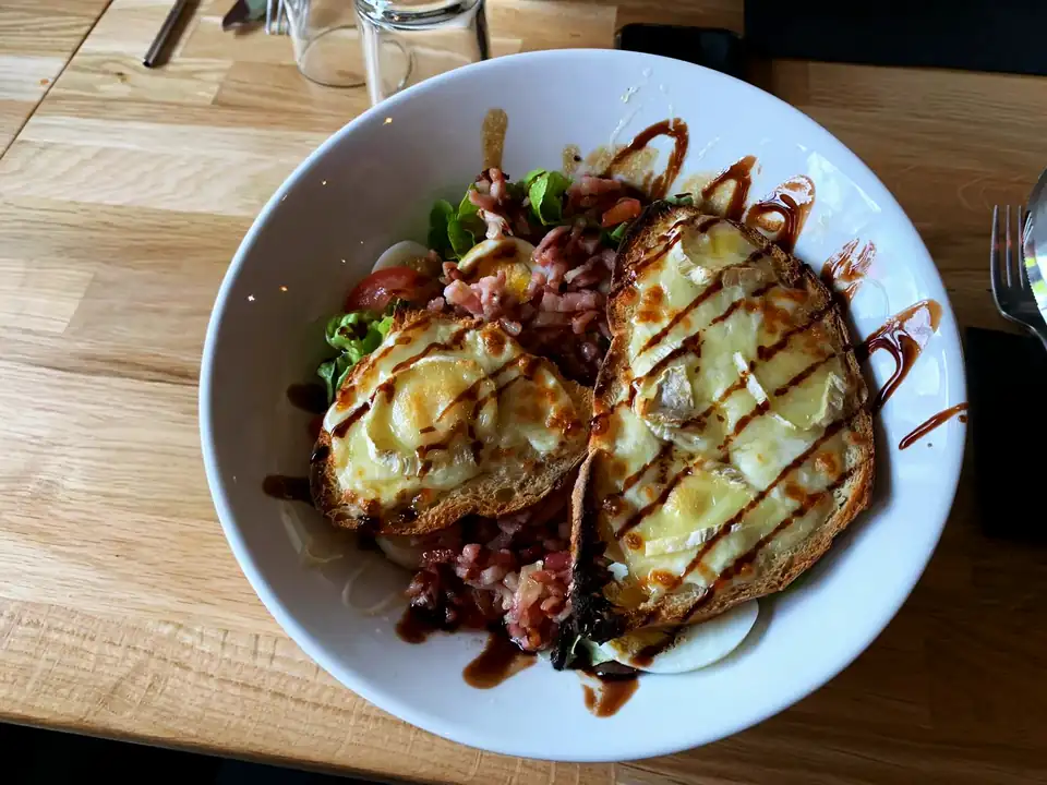 Une salade Pili servie en salle : tartines de chèvre et de camembert gratinées, lardons, œuf, tomate et glaçage balsamique.