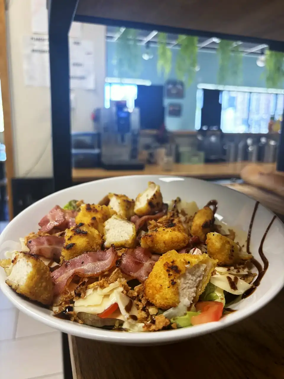Une salade au chèvre pané, bacon et oignons frits posée sur le comptoir, avec la salle de Pili Pizza et son mur vert en arrière-plan.