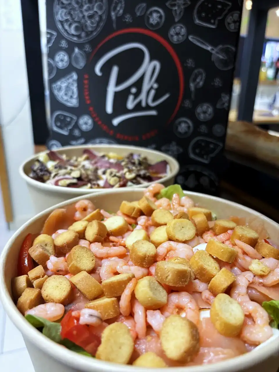 Deux salades à emporter en bols kraft, l'une aux crevettes et croûtons, l'autre au pastrami, devant une boîte à pizza Pili.