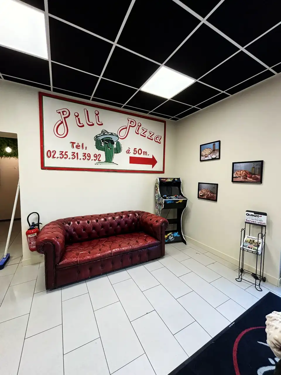 Le salon d'attente de Pili Pizza : l'enseigne peinte d'origine avec un cactus au sombrero et le numéro de téléphone, un canapé Chesterfield bordeaux et une borne d'arcade.