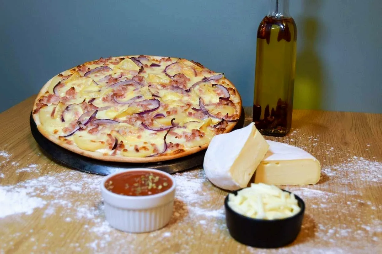 Une pizza Pili à la pomme de terre, aux lardons et à l'oignon rouge sur une table en bois fariné, à côté d'un reblochon entamé, d'un ramequin de mozzarella et d'une huile pimentée.